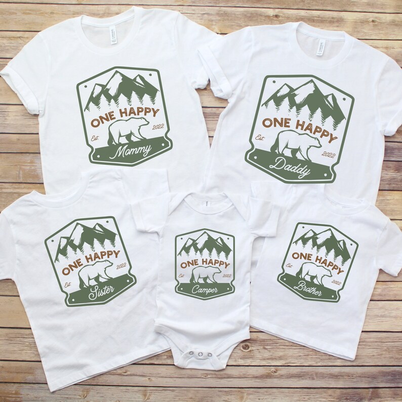 May include: Set of five white t-shirts with a green mountain and bear design. The shirts say "One Happy" with the year 2022 and the words "Mommy", "Daddy", "Sister", "Camper", and "Brother".