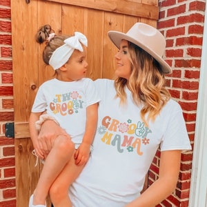 May include: A white t-shirt with the text "Groovy Mama" in colorful retro lettering and flowers. A child is wearing a white t-shirt with the text "Two Groovy" in colorful retro lettering and flowers.