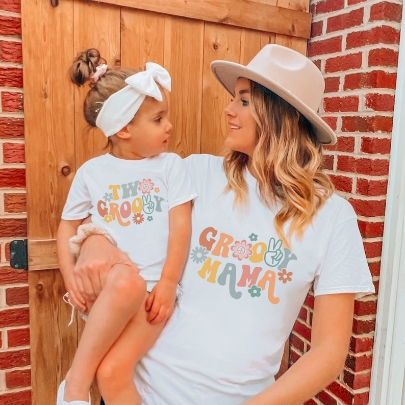 May include: A white t-shirt with the text "Groovy Mama" in colorful retro lettering and flowers. A child is wearing a white t-shirt with the text "Two Groovy" in colorful retro lettering and flowers.