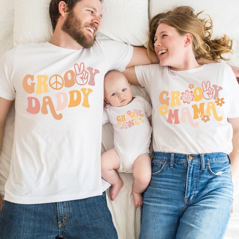 May include: A family of three wearing matching white t-shirts with a groovy font design. The shirts say "Groovy Daddy", "Groovy Mama", and "Groovy One".