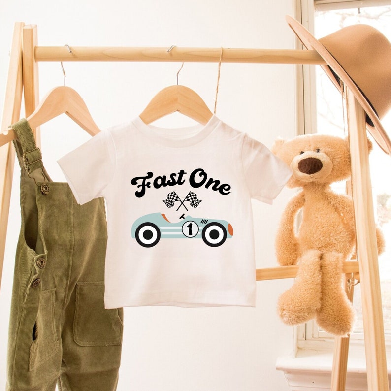 May include: White baby onesie with a blue race car graphic and the text "Fast One" in black. The race car has a number one on the side and checkered flags.