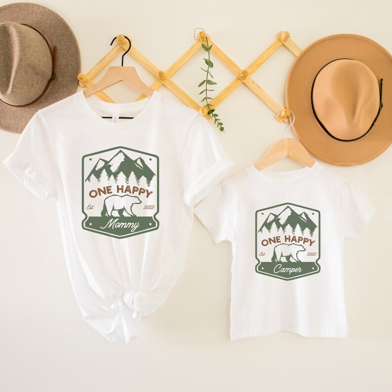 May include: White t-shirts with a green mountain and bear graphic. The larger shirt says "One Happy Mommy Est. 2022" and the smaller shirt says "One Happy Camper Est. 2022".