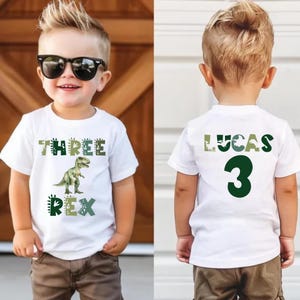 May include: A white t-shirt with a dinosaur graphic and the words "THREE REX" in green and gray. The back of the shirt has the name "LUCAS" and the number "3" in green. The shirt is worn by a young child.