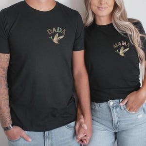 May include: A couple wearing black t-shirts with the words "DADA" and "MAMA" and a flying duck graphic. The shirts are a dark charcoal colour.
