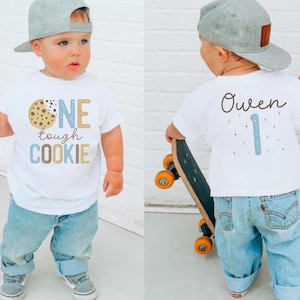 Peut inclure: T-shirt blanc avec l'inscription "ONE tough COOKIE" et un motif de biscuit. Le dos du t-shirt porte le nom "Owen" et le chiffre "1". L'enfant porte un jean bleu et une casquette de baseball grise.