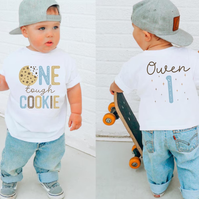 May include: A white t-shirt with the text "ONE tough COOKIE" and a cookie graphic. The back of the shirt has the name "Owen" and the number "1". The child is wearing blue jeans and a gray baseball cap.