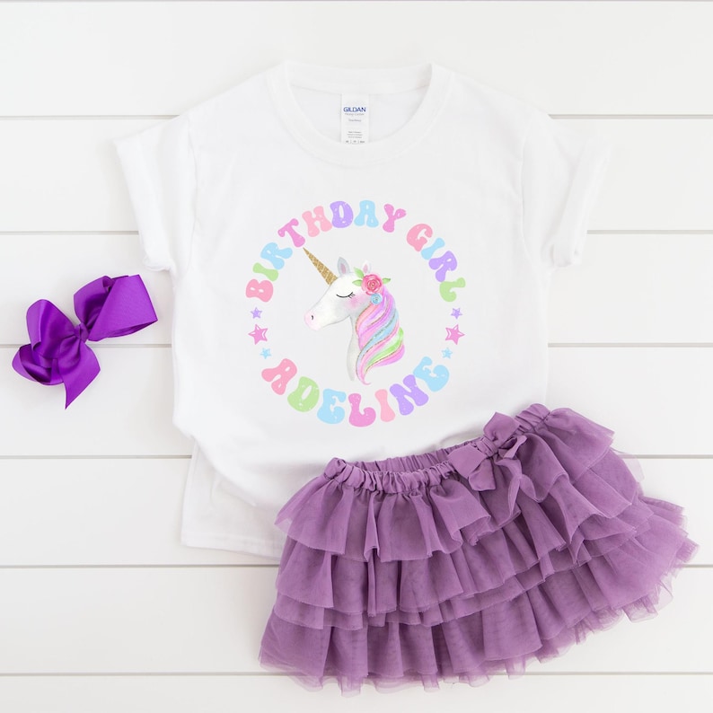 Puede incluir: Una camiseta blanca con un gr&aacute;fico de unicornio colorido y el texto "Birthday Girl Adeline". Se combina con una falda tut&uacute; morada y un lazo a juego. Un atuendo festivo para una celebraci&oacute;n de cumplea&ntilde;os.