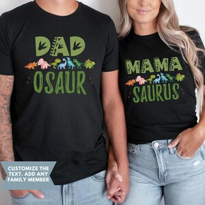 May include: Two black t-shirts with a dinosaur theme. The first shirt says "Dad-osaur" with a green dinosaur footprint design. The second shirt says "Mama-saurus" with a green dinosaur footprint design.