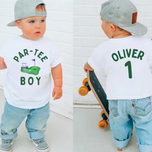 May include: A white t-shirt with the text "PAR-TEE BOY" and a golf-themed graphic. The back of the shirt has the name "OLIVER" and the number "1" in green. The child is wearing blue jeans and a gray baseball cap.