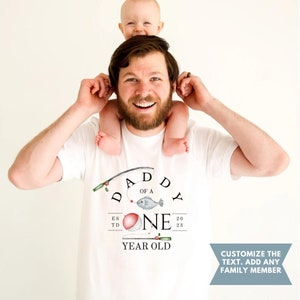 May include: White t-shirt with a fishing rod graphic and the text "Daddy of a One Year Old" with a red and white fishing bobber.