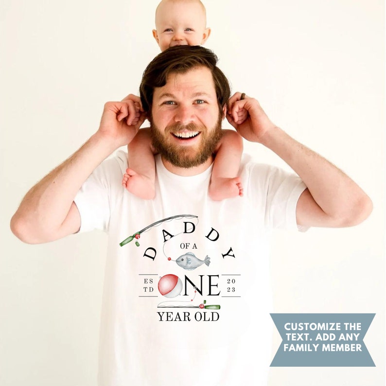 May include: White t-shirt with a fishing rod graphic and the text "Daddy of a One Year Old" with a red and white fishing bobber.