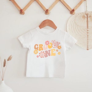 May include: White t-shirt with a groovy one graphic in retro colours with flowers and a peace sign.