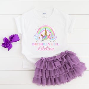 K&ouml;nnte beinhalten: Wei&szlig;es T-Shirt mit einem Einhorn- und Regenbogen-Design. Der Text "Birthday Girl Adeline" ist auf dem Shirt aufgedruckt. Ein lilafarbener T&uuml;llrock ist ebenfalls abgebildet.