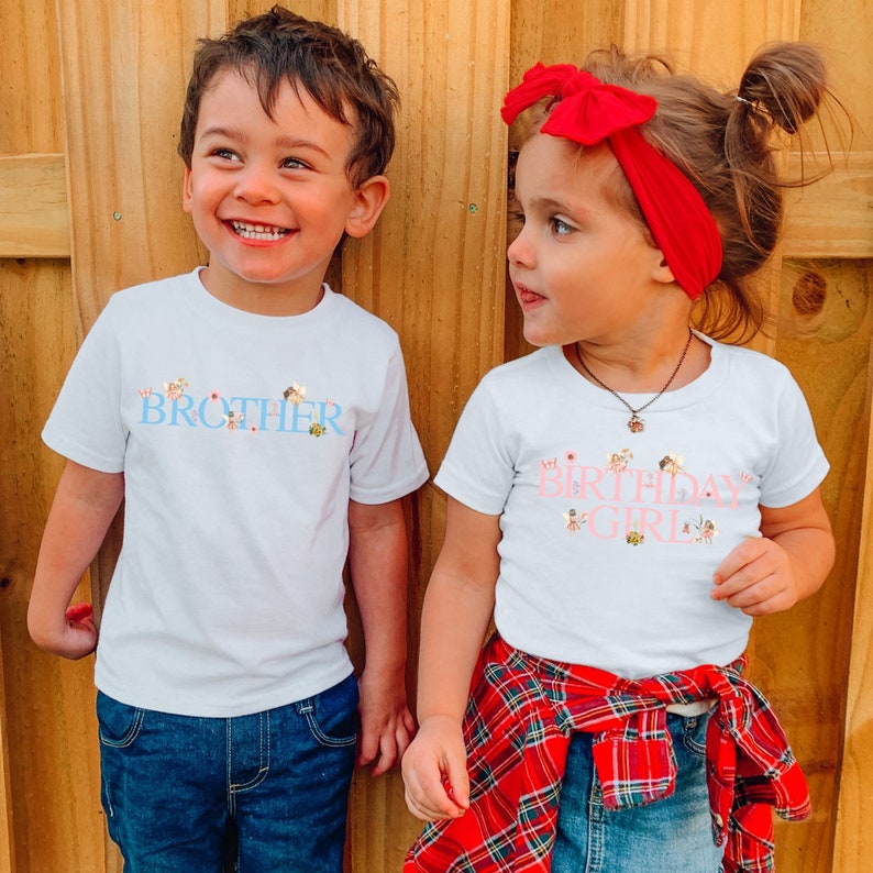 May include: Two white short-sleeved t-shirts. The left shirt features the word BROTHER in blue letters with small fairy graphics. The right shirt features the words BIRTHDAY GIRL in pink letters with small fairy graphics. Both shirts have crew necks.