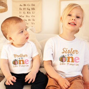 Puede incluir: Dos ni&ntilde;os con camisas blancas con un gr&aacute;fico de calabaza y el texto "Brother of the One Little Pumpkin" y "Sister of the One Little Pumpkin".