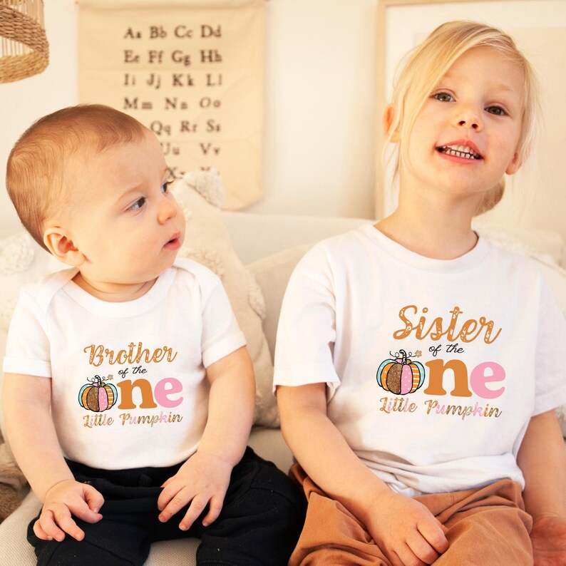 Puede incluir: Dos ni&ntilde;os con camisas blancas con un gr&aacute;fico de calabaza y el texto "Brother of the One Little Pumpkin" y "Sister of the One Little Pumpkin".