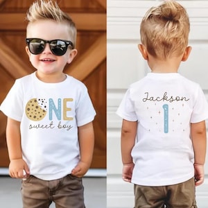Peut inclure: Un t-shirt blanc avec un biscuit et les mots "ONE sweet boy" en bleu et or. Le dos du t-shirt porte le nom "Jackson" et le chiffre 1 bleu. Le garçon porte des lunettes de soleil et un pantalon marron.