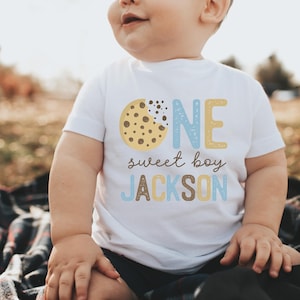May include: A white t-shirt with the words "ONE sweet boy JACKSON" printed on the front. The "ONE" is in blue and yellow, and the "sweet boy" and "JACKSON" are in brown and blue. A cookie graphic is on the left.