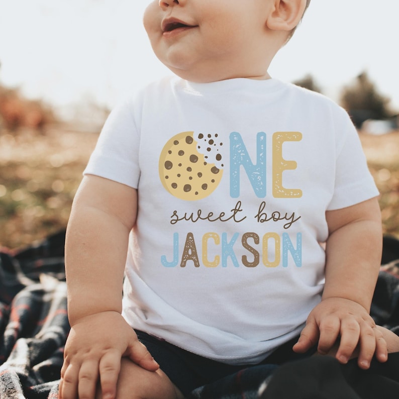 May include: A white t-shirt with the words "ONE sweet boy JACKSON" printed on the front. The "ONE" is in blue and yellow, and the "sweet boy" and "JACKSON" are in brown and blue. A cookie graphic is on the left.