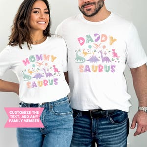 K&ouml;nnte beinhalten: Zwei wei&szlig;e T-Shirts mit bunten Dinosaurier-Grafiken und den Worten "Mommy Saurus" und "Daddy Saurus". Die Shirts haben ein verspieltes Design mit Pastellfarben. Das Bild enth&auml;lt auch den Text "Customize the text, add any family member".