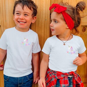 Puede incluir: Dos ni&ntilde;os con camisetas blancas con un gr&aacute;fico de unicornio y la palabra "Sister" en colores pastel. La ni&ntilde;a tiene un lazo rojo en el pelo y una camisa a cuadros roja y negra atada a la cintura.