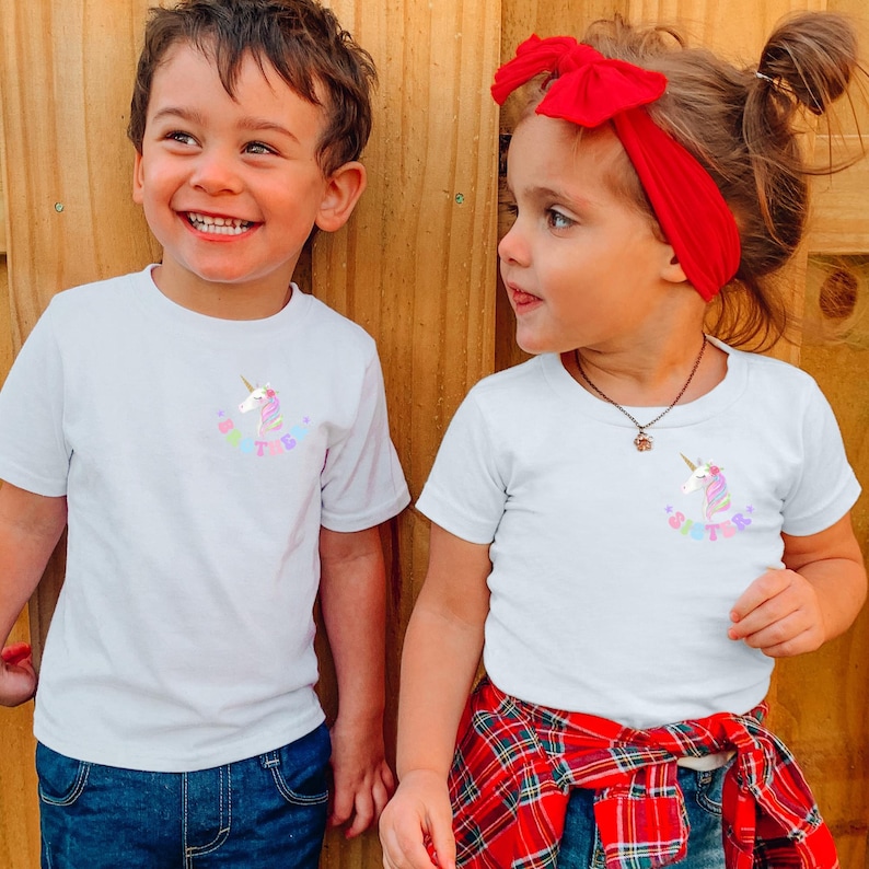 Puede incluir: Dos ni&ntilde;os con camisetas blancas con un gr&aacute;fico de unicornio y la palabra "Sister" en colores pastel. La ni&ntilde;a tiene un lazo rojo en el pelo y una camisa a cuadros roja y negra atada a la cintura.