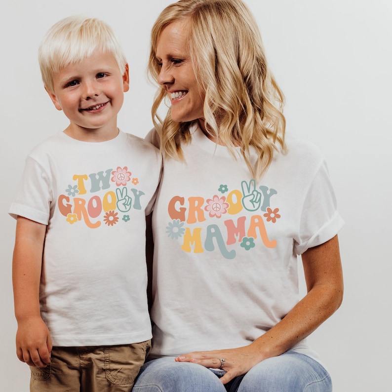 May include: A white t-shirt with the text "Two Groovy" in colorful letters and a peace sign. A second white t-shirt with the text "Groovy Mama" in colorful letters and a peace sign. The shirts are worn by a child and an adult.