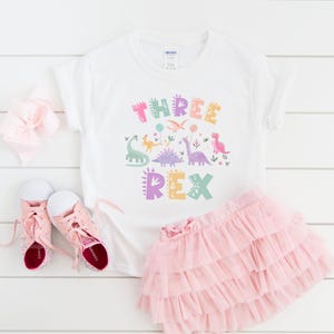K&ouml;nnte beinhalten: Wei&szlig;es T-Shirt mit einem bunten "THREE REX" Dinosaurier-Motiv, kombiniert mit einem rosa Tutu-Rock und rosa Turnschuhen. Eine rosa Schleife ist ebenfalls im Bild. Das Outfit ist f&uuml;r ein Kind konzipiert.