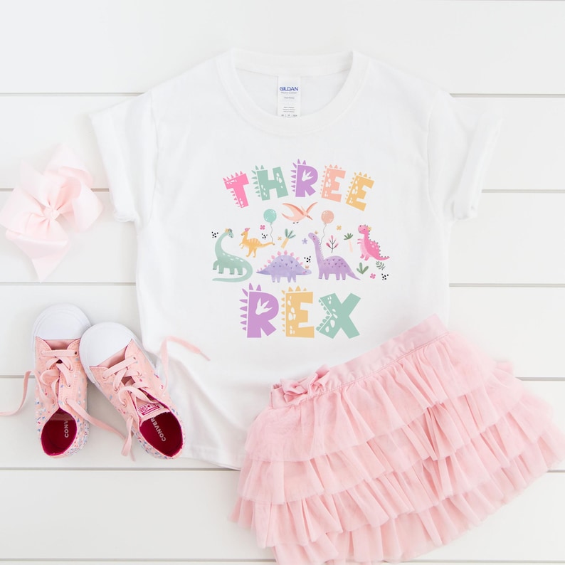 K&ouml;nnte beinhalten: Wei&szlig;es T-Shirt mit einem bunten "THREE REX" Dinosaurier-Motiv, kombiniert mit einem rosa Tutu-Rock und rosa Turnschuhen. Eine rosa Schleife ist ebenfalls im Bild. Das Outfit ist f&uuml;r ein Kind konzipiert.