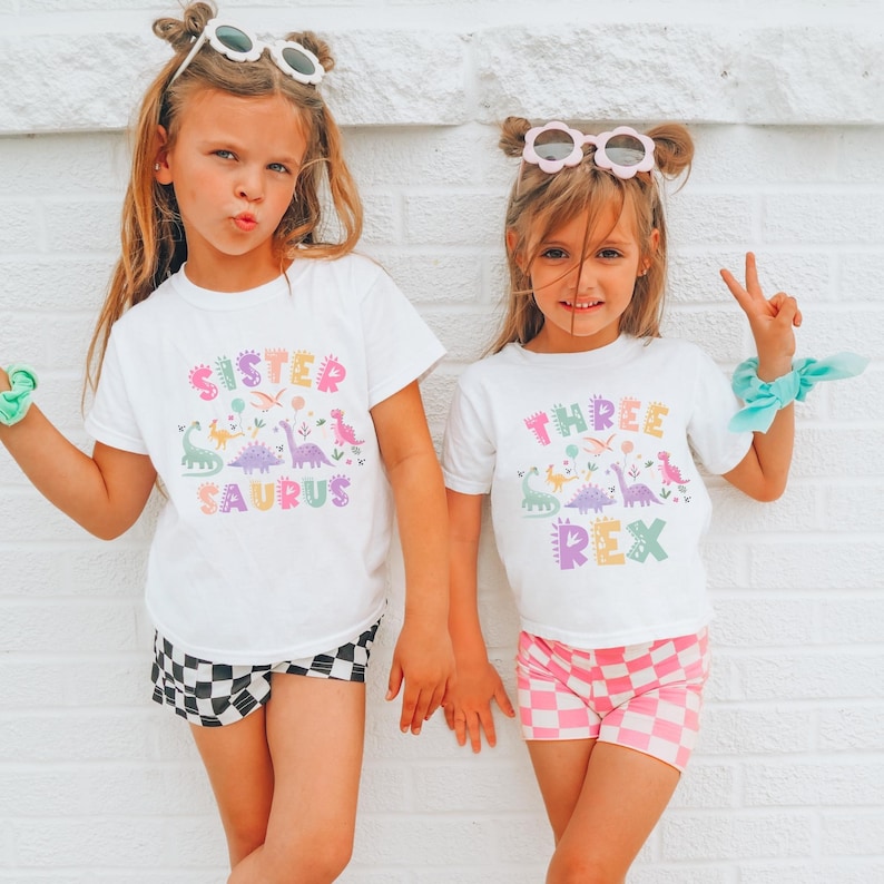 K&ouml;nnte beinhalten: Zwei junge M&auml;dchen in passenden wei&szlig;en T-Shirts mit Dinosaurier-Grafiken und Text. Ein Shirt tr&auml;gt die Aufschrift "Sister Saurus" und das andere "Three Rex". Die M&auml;dchen tragen au&szlig;erdem Shorts, eine mit einem schwarz-wei&szlig;en Schachbrettmuster und die andere mit einem rosa-wei&szlig;en Schachbrettmuster.