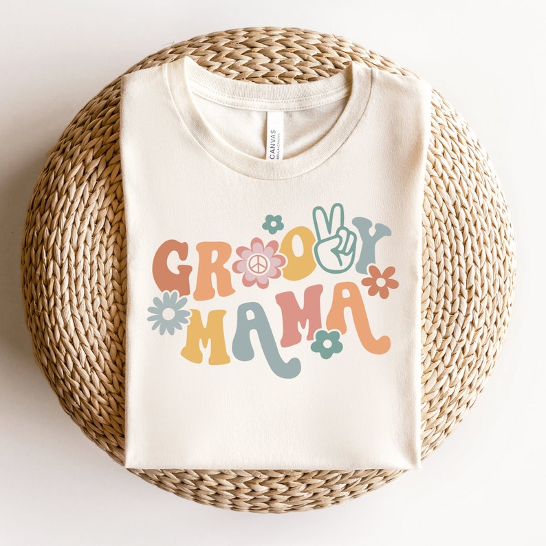 May include: A white t-shirt with the text "Groovy Mama" in colorful retro lettering. The text is surrounded by peace signs and flowers.