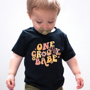 Może przedstawiać: Czarny T-shirt z kr&oacute;tkim rękawem z kolorową grafiką, na kt&oacute;rej widnieje napis "ONE GROOVY BABE" w retro czcionce. Grafika zawiera symbole pokoju i kwiaty.