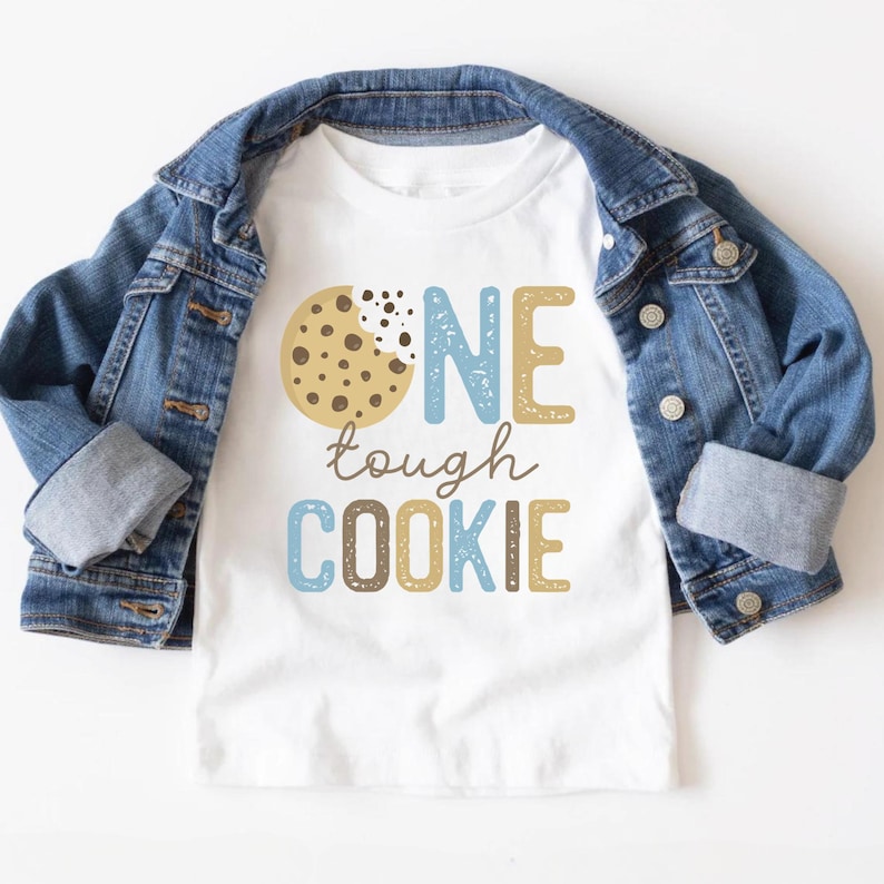 May include: White t-shirt with the text "ONE tough COOKIE" in blue, brown, and tan colors. A denim jacket is draped over the shirt. The design includes a graphic of a cookie with a bite taken out of it.