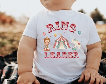 Puede incluir: Camiseta blanca de manga corta con un diseño de circo. El diseño incluye las palabras "RING LEADER" en rojo, junto con ilustraciones de una carpa de circo, un elefante y un león. La camiseta la lleva un bebé.