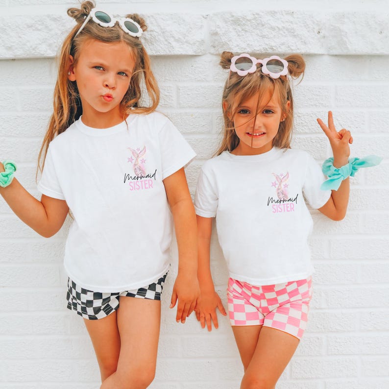 May include: Two young girls wearing matching white t-shirts with a pink mermaid graphic and the text "Mermaid Sister". One girl wears black and white checkered shorts, the other wears pink and white checkered shorts. Both wear sunglasses and scrunchies.