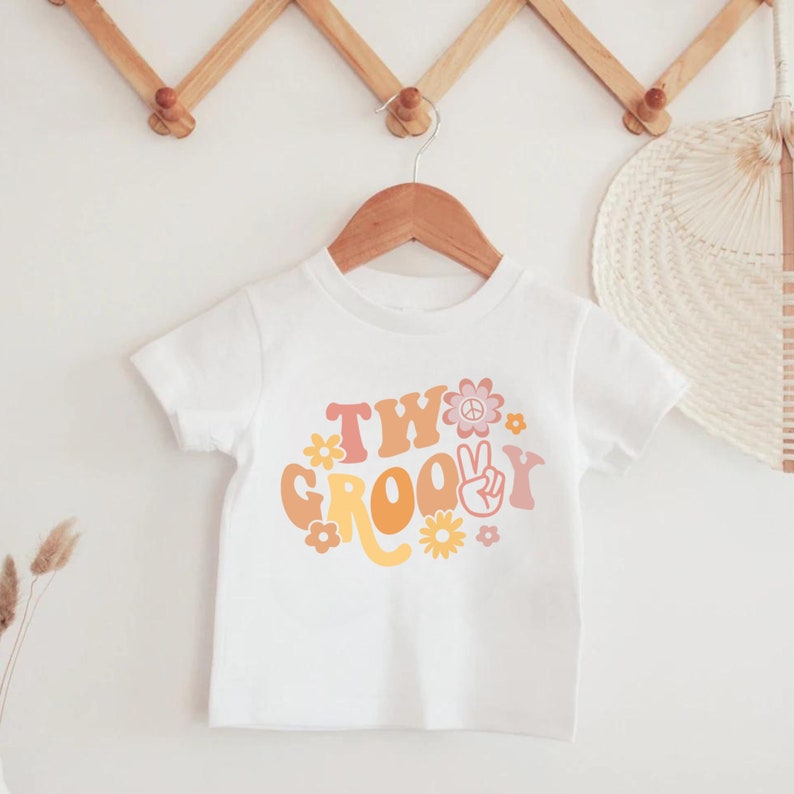 May include: White t-shirt with a graphic design that says "Two Groovy" in a retro font with peace signs and flowers.