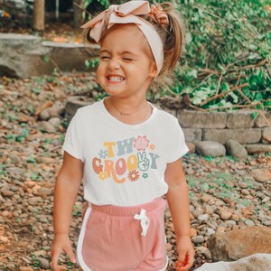 May include: A young child wearing a white t-shirt with the text "Two Groovy" in colorful retro font and a peace sign graphic. The child is wearing pink shorts and a pink headband.