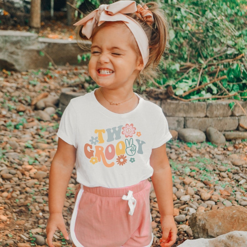May include: A young child wearing a white t-shirt with the text "Two Groovy" in colorful retro font and a peace sign graphic. The child is wearing pink shorts and a pink headband.