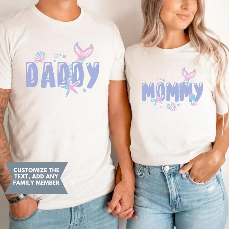 May include: Two white t-shirts with a mermaid tail design and the words "Daddy" and "Mommy" in pastel colors. The shirts are worn by two people holding hands.