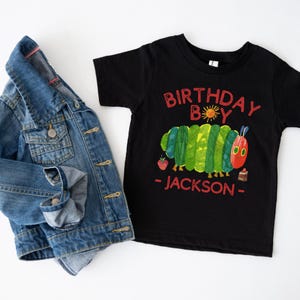 Może przedstawiać: Czarny t-shirt z kolorową grafiką gąsienicy i napisem "BIRTHDAY BOY - JACKSON -". Obok koszulki leży niebieska kurtka dżinsowa. Koszulka ma czerwony, zielony i ż&oacute;łty wz&oacute;r.