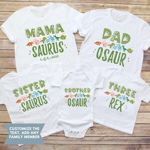 May include: Set of five white t-shirts with dinosaur graphics and text. The shirts say "Mama Saurus est. 2023", "Dad Osaur", "Sister Saurus", "Brother Osaur", and "Three Rex".