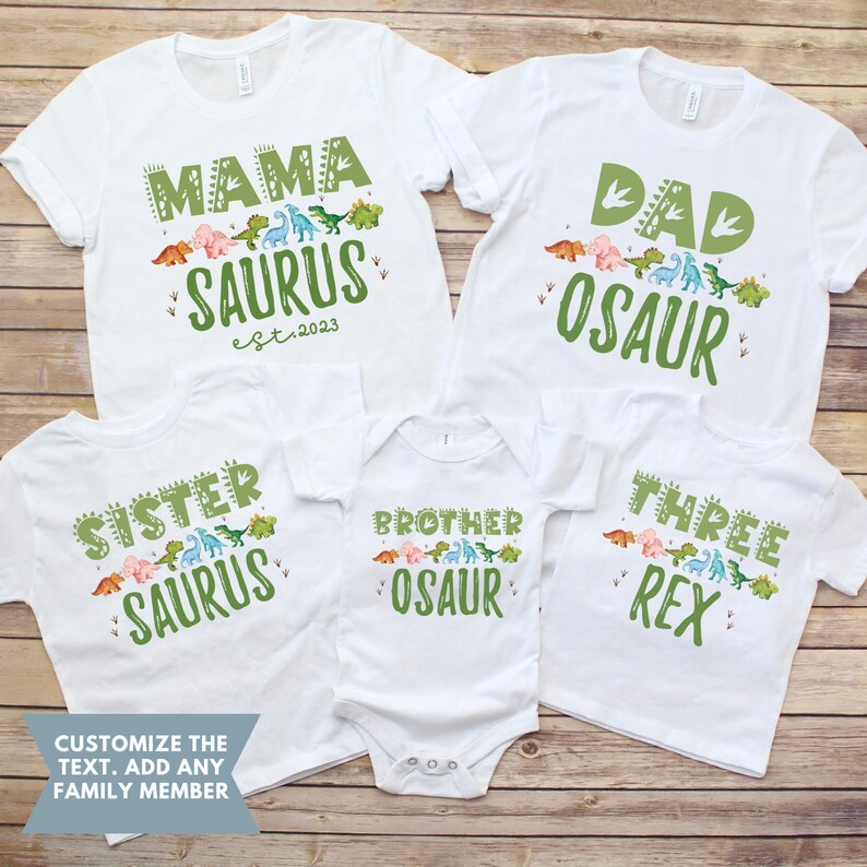 May include: Set of five white t-shirts with dinosaur graphics and text. The shirts say "Mama Saurus est. 2023", "Dad Osaur", "Sister Saurus", "Brother Osaur", and "Three Rex".