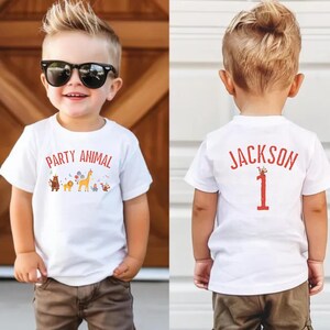 Peut inclure: Un jeune enfant portant un t-shirt blanc avec les mots "PARTY ANIMAL" et un graphisme animalier color&eacute;. L'enfant porte &eacute;galement des lunettes de soleil et un pantalon marron. Le dos du t-shirt porte le nom "JACKSON" et le chiffre "1".
