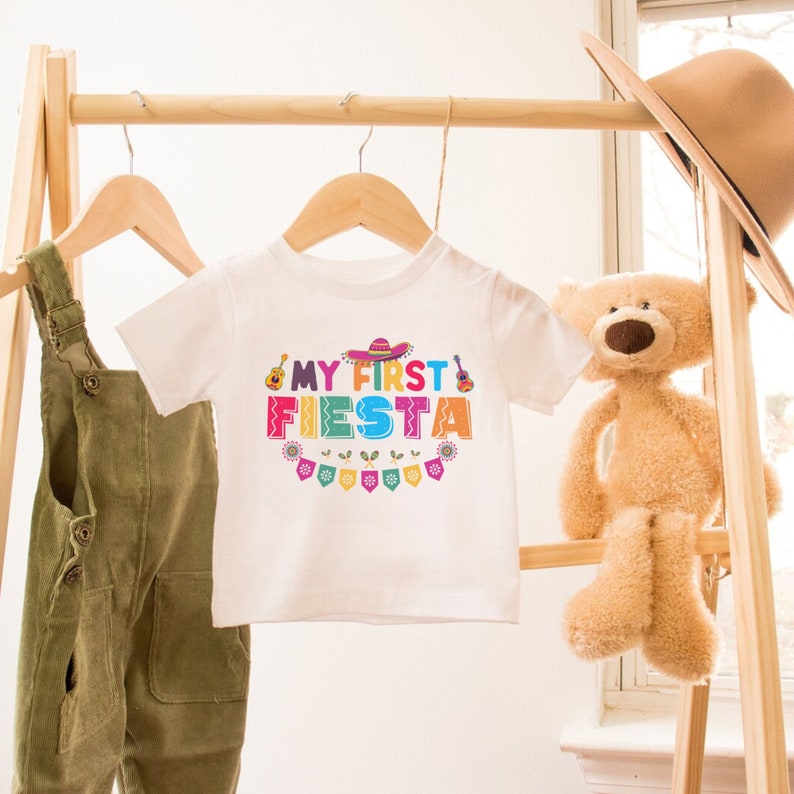 K&ouml;nnte beinhalten: Ein wei&szlig;er Baby-Strampler mit dem Text "My First Fiesta" in bunten Buchstaben mit einem Sombrero, einer Gitarre und festlichen Flaggen.