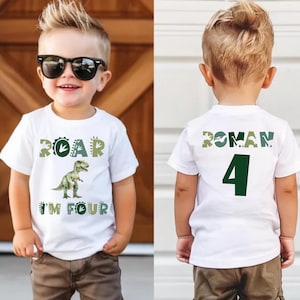 May include: White t-shirt with a green dinosaur graphic and the text "ROAR I'M FOUR". The back of the shirt has the name "ROMAN" and the number "4" in green. The child is wearing sunglasses and brown pants.