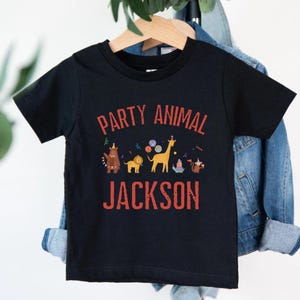 Peut inclure: T-shirt noir pour enfant avec l'inscription "PARTY ANIMAL JACKSON" en rouge. Le t-shirt pr&eacute;sente un graphisme color&eacute; d'animaux de dessins anim&eacute;s, dont une girafe, un lion et un dinosaure, avec des d&eacute;corations de f&ecirc;te.