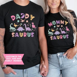 K&ouml;nnte beinhalten: Schwarze T-Shirts mit bunten Dinosaurier-Grafiken und den Worten "Daddy Saurus" und "Mommy Saurus". Die Shirts haben verschiedene Dinosaurier-Illustrationen und den Text "Customize the text. Add any family member" in einem rosa Banner.