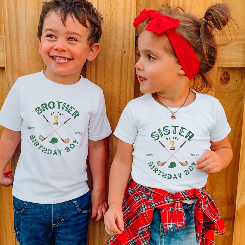 May include: Two children wearing white t-shirts with green and gold golf-themed graphics. The shirts say "Brother of the Birthday Boy" and "Sister of the Birthday Boy".