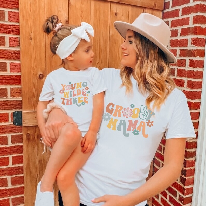 May include: A mother and daughter wearing matching white t-shirts. The daughter's shirt says "Young Wild & Three" with flowers and peace signs. The mother's shirt says "Groovy Mama" with flowers and peace signs.