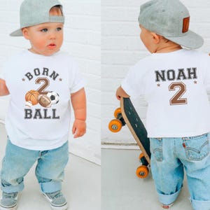 Peut inclure: T-shirt blanc avec l'inscription "BORN 2 BALL" et des graphiques de ballons de sport. Le dos du t-shirt porte le nom "NOAH" et le chiffre "2". L'enfant porte un jean bleu et une casquette de baseball grise.